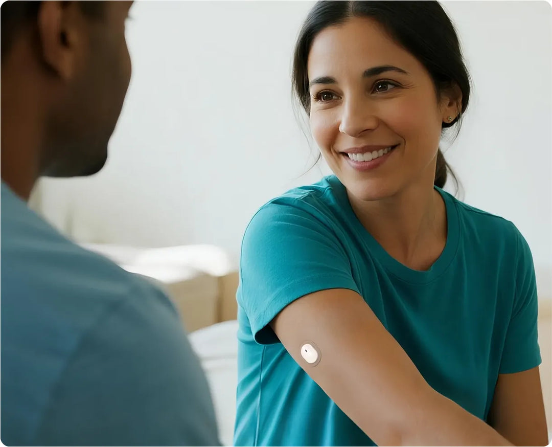 A person wearing a teal shirt with a SIBIONICS glcose monitor patch on their arm, interacting with another individual. 